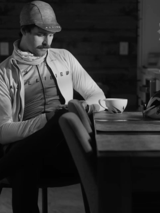 Person sitting at a table with a cup, wearing a jacket with visible branding, in a dimly lit room in black and white