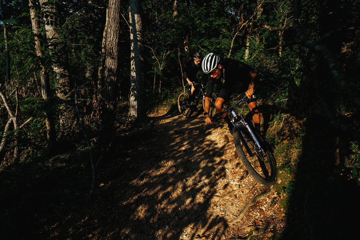 Two mountain bikers riding ALLIED BC40s on a forest trail at sunset, with one rider in the foreground wearing a white helmet and black clothing.